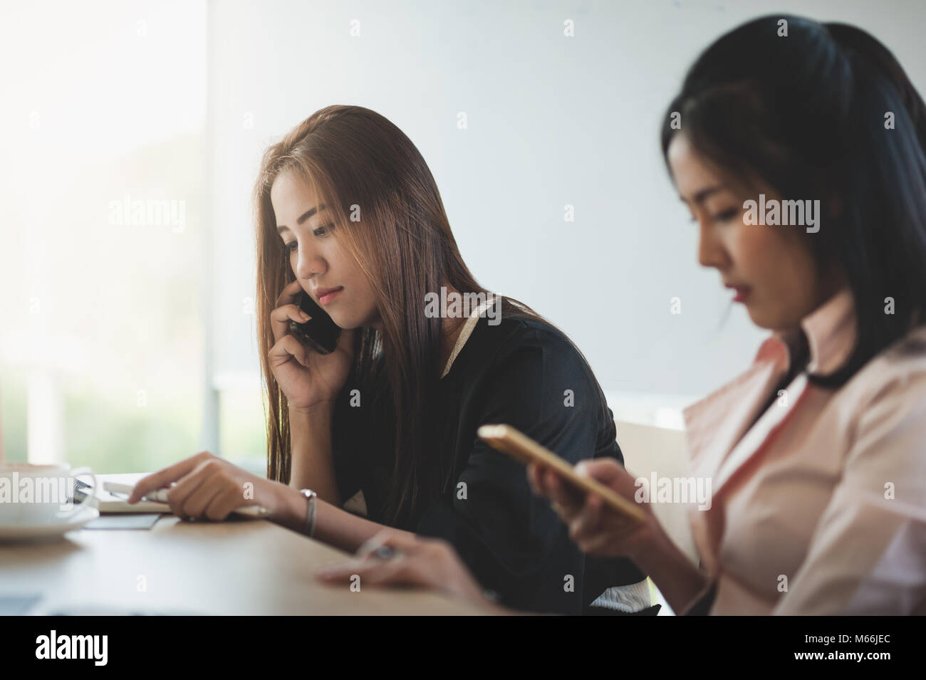 Young Asian businesswoman talking on smartphone while sitting beside colleague on break time in office. Wireless communication concept Stock Photo