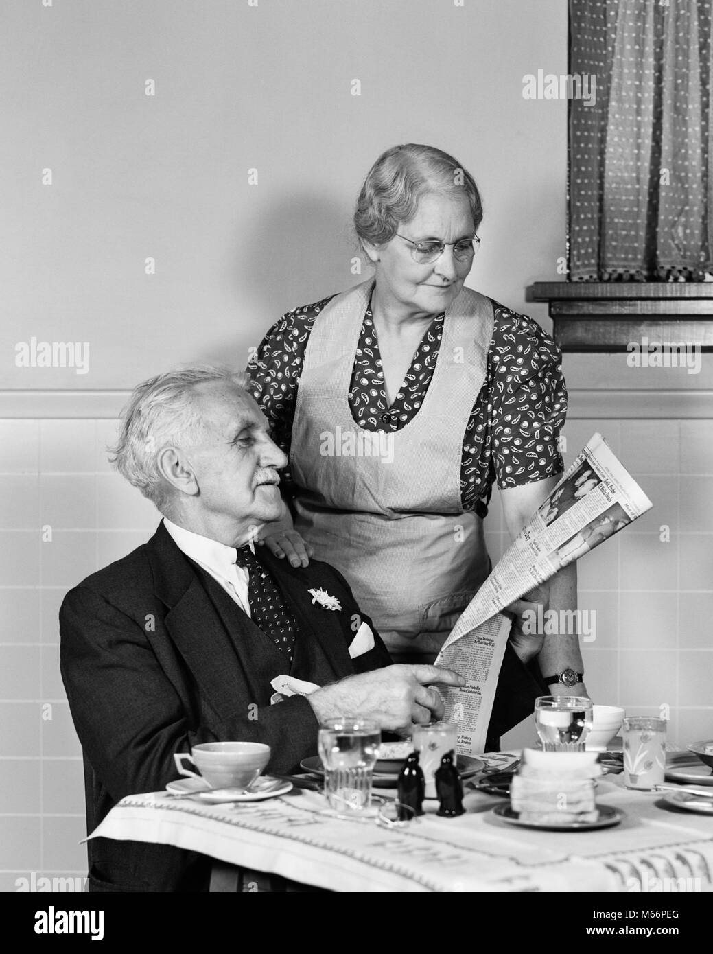 1940s ELDERLY COUPLE MAN WOMAN IN KITCHEN MAN SITTING AT TABLE WOMAN LOOKING OVER HIS SHOULDER READING NEWSPAPER - r13244 HAR001 HARS CAUCASIAN PLEASED JOY ELDER FEMALES MARRIED STUDIO SHOT SPOUSE HUSBANDS HEALTHINESS HOME LIFE COPY SPACE FRIENDSHIP HALF-LENGTH LADIES CARING NEWSPAPERS COUPLES INDOORS RETIREMENT SENIOR MAN SENIOR ADULT NOSTALGIA MORNING TOGETHERNESS SENIOR WOMAN HOMEMAKER WIVES HOMEMAKERS OLDSTERS CHEERFUL OLDSTER HIS HOUSEWIVES SMILES ELDERS JOYFUL 70s ADULT ELDERLY MAN 80-PLUS ADULT ELDERLY WOMAN MALES B&W BLACK AND WHITE CAUCASIAN ETHNICITY HOME MAKER HOUSE WIFE HOUSEKEEPER Stock Photo