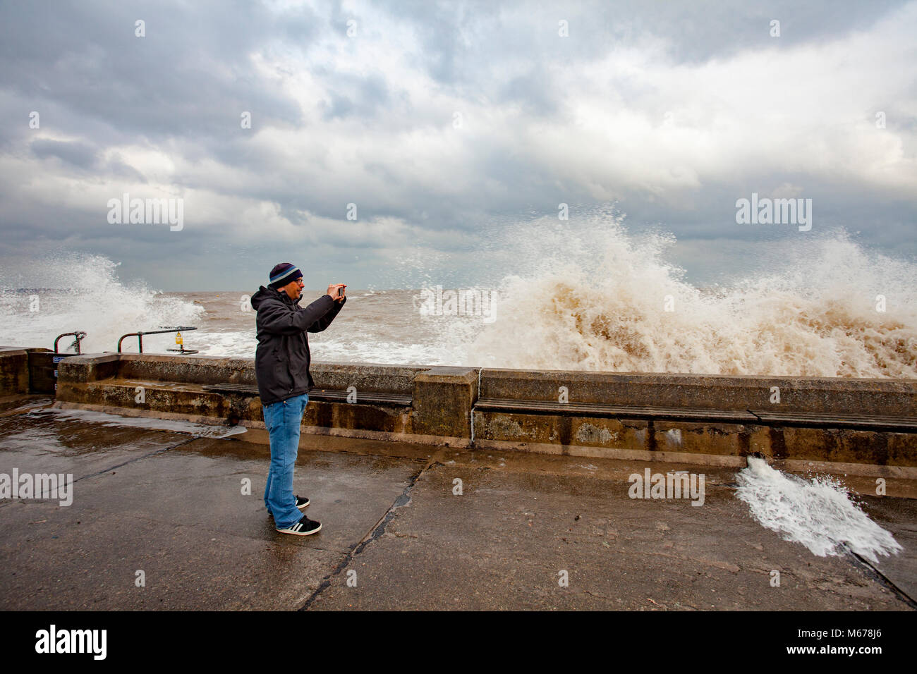 A man taking pictures of the crashing waves on the promenade wall at Colwyn Bay, Wales as a result of the Beast from the East and Storm Emma creating huge destructive waves along the coastline Stock Photo