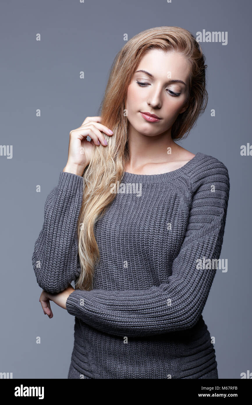 Portrait of young woman in gray woolen sweater. Beautiful girl posing on grey studio background. Female with blonde hair and day beauty makeup. Stock Photo