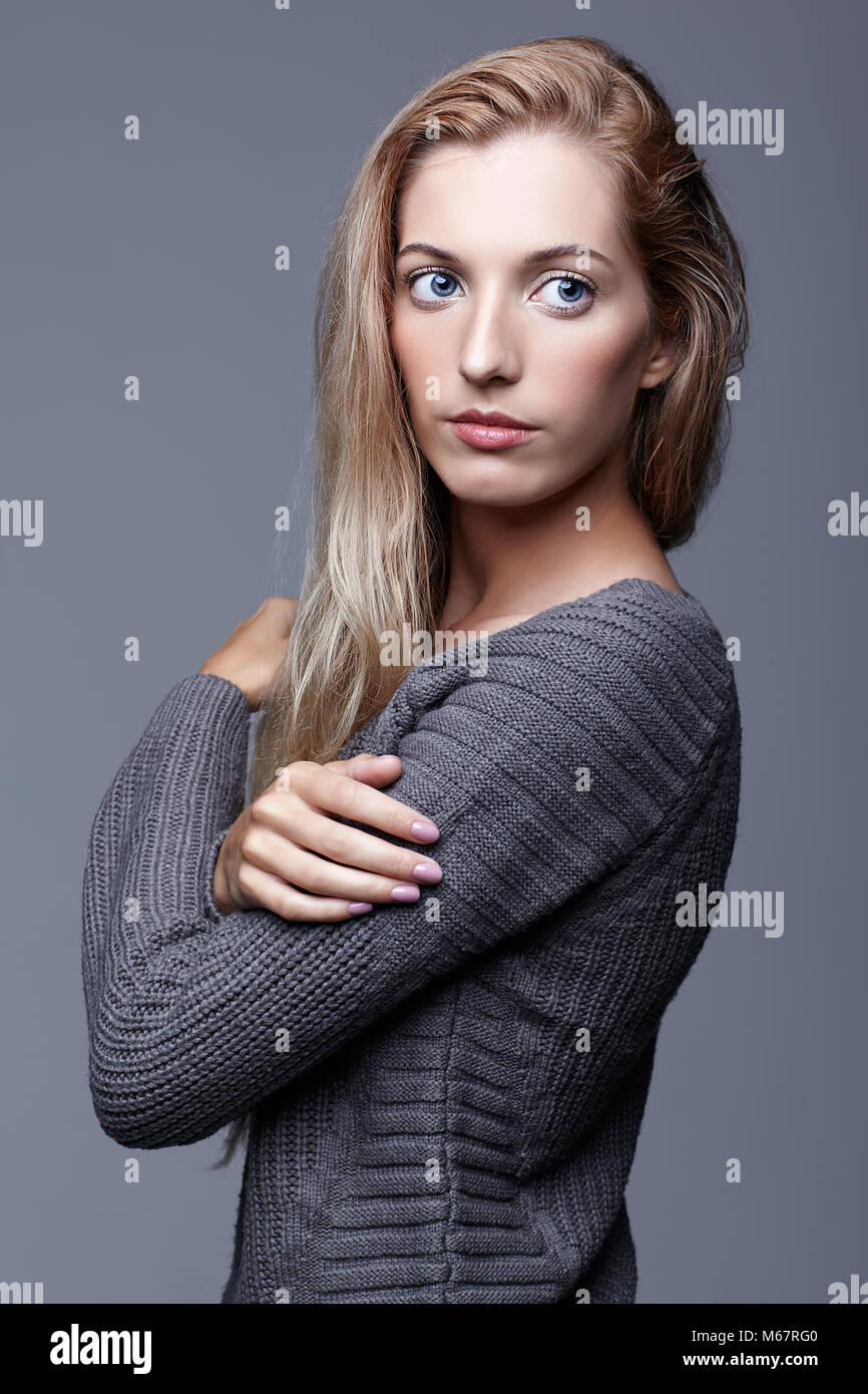Portrait of young woman in gray woolen sweater. Beautiful girl posing on grey studio background. Female with blonde hair and day beauty makeup. Stock Photo