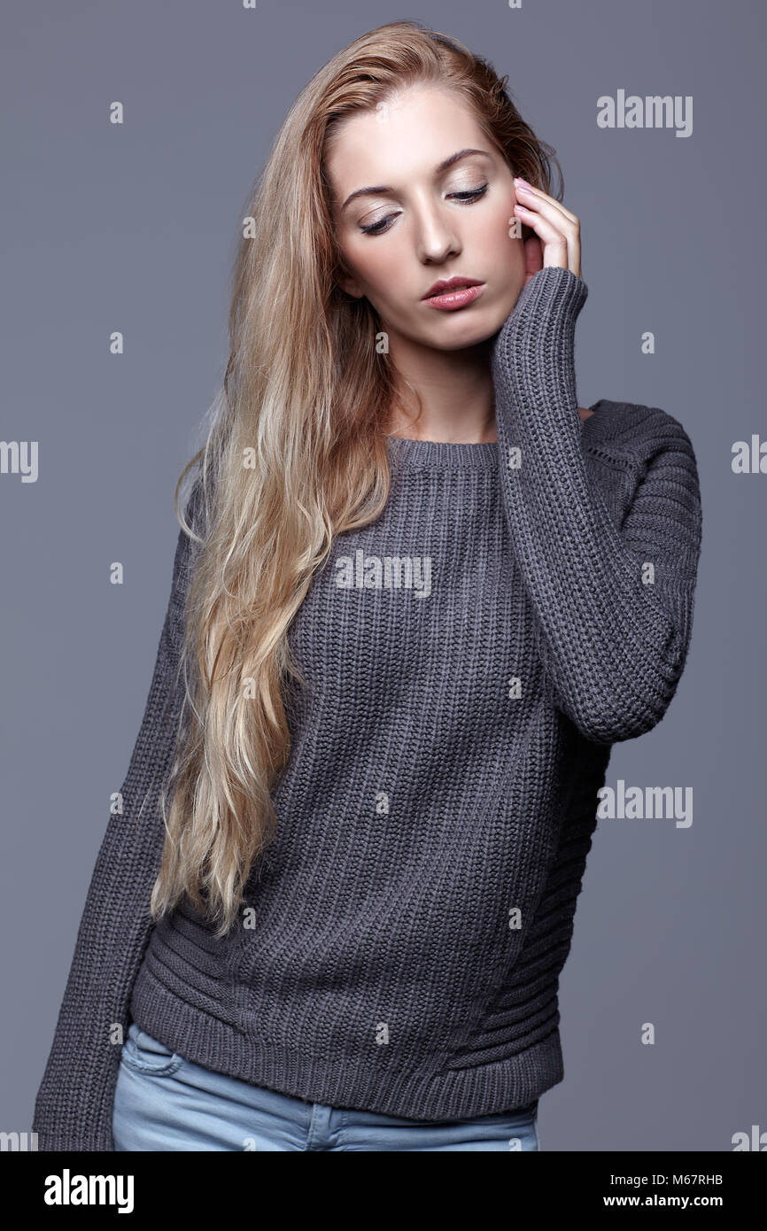 Portrait of young woman in gray woolen sweater and jeans. Beautiful girl posing on grey studio background. Female with blonde hair and day beauty make Stock Photo