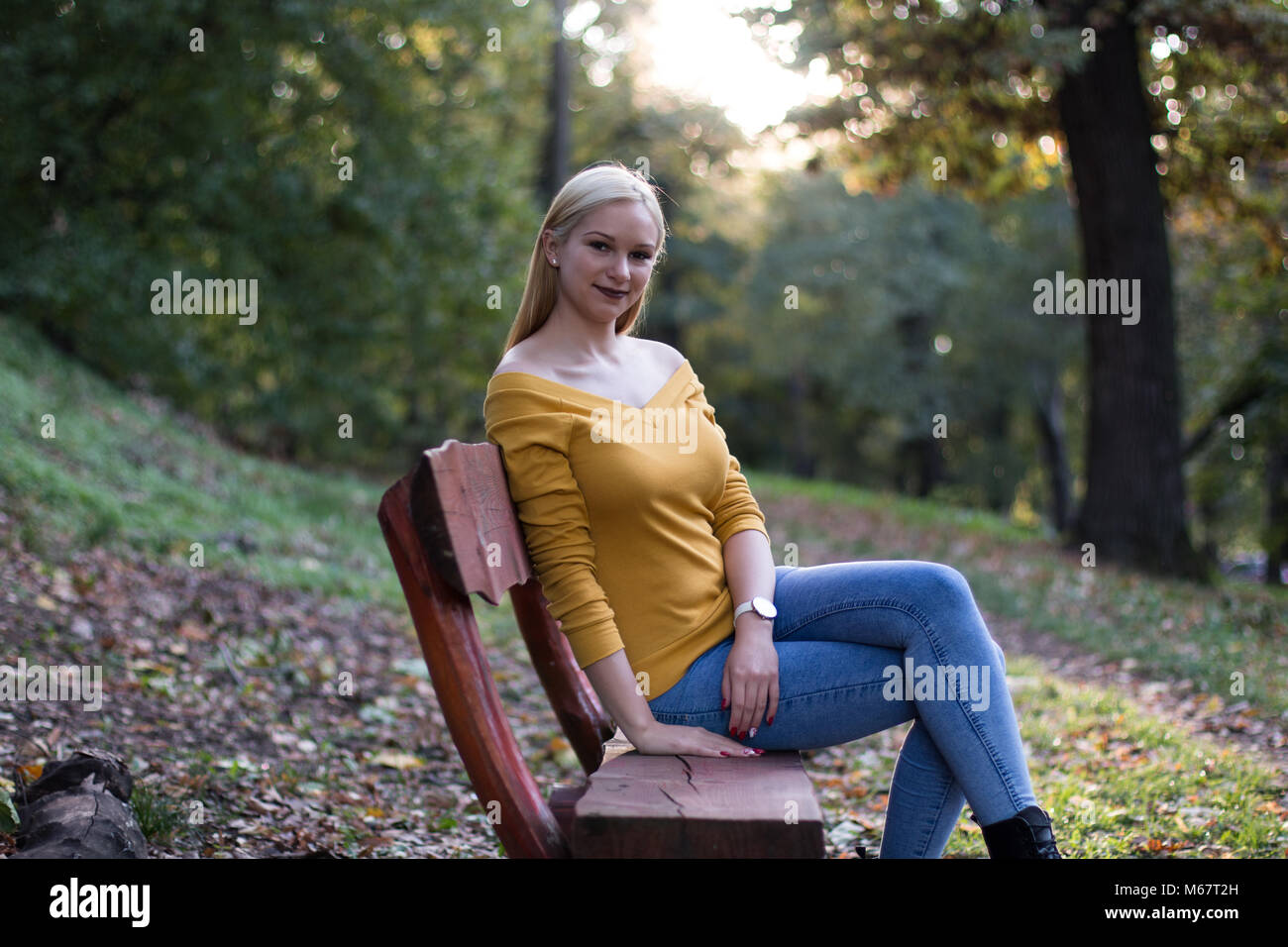 Young beautiful blonde woman sitting on a wooden bench in the park ...