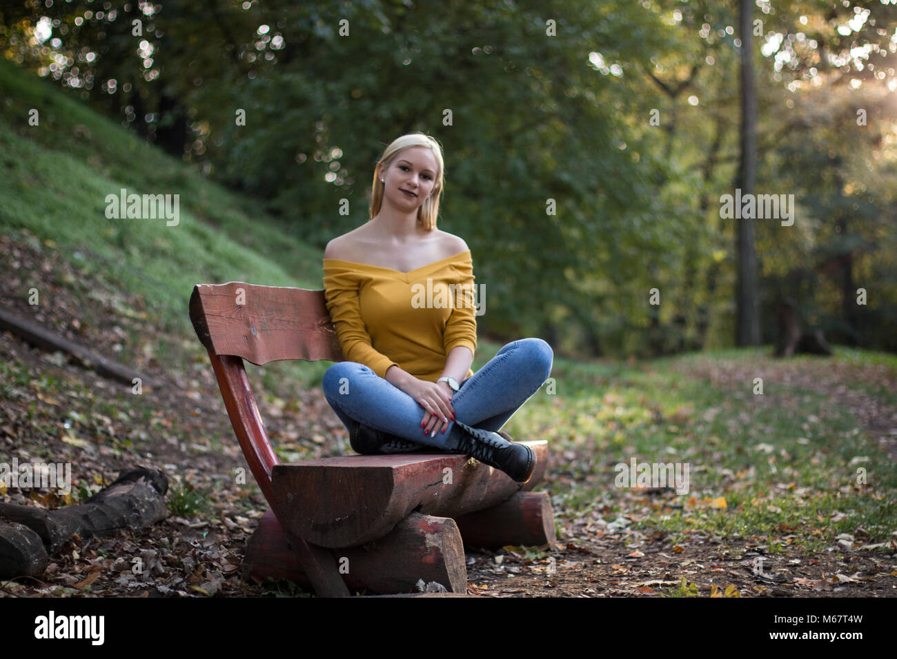 Young beautiful blonde woman sitting on a wooden bench in the park ...