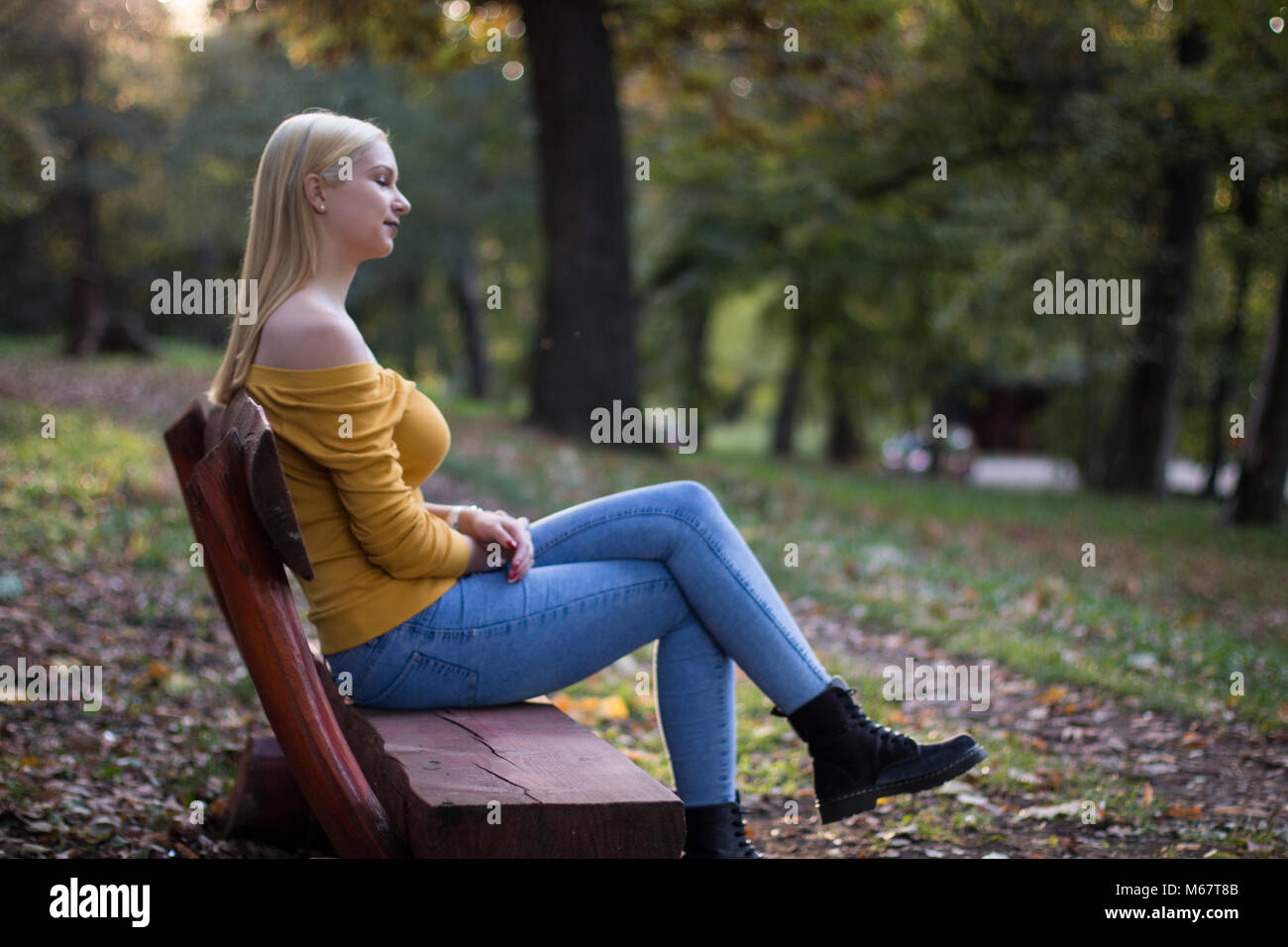 Young beautiful blonde woman sitting on a wooden bench in the park ...