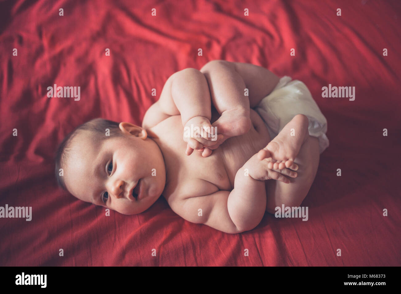 A cute little baby in his diaper is lying on a red bed sheet Stock Photo