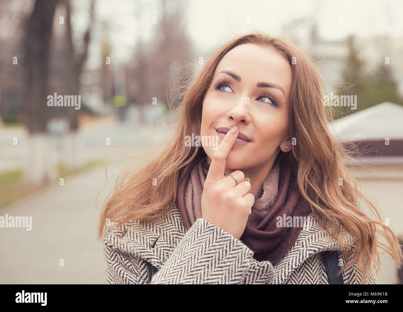Young pretty woman in coat touching lips and daydreaming while standing on street and looking away. Stock Photo