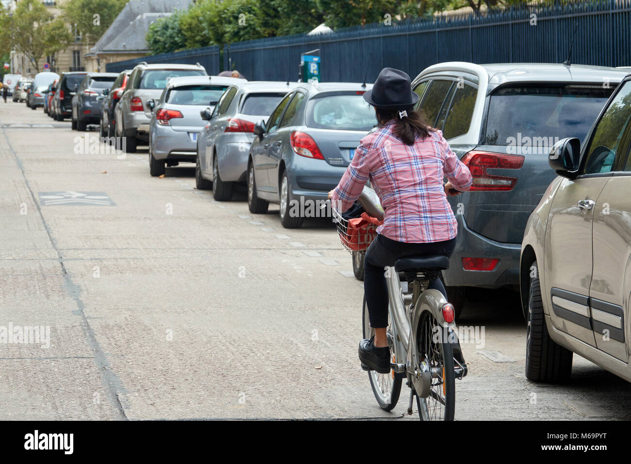 A woman in city clothes in a hat rides a bicycle along parked cars on a Paris street. Stock Photo