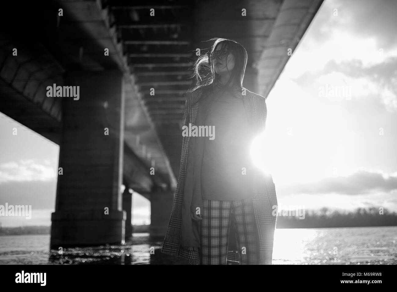 Young woman in checkered coat walks under bridge at sunset. Black and ...
