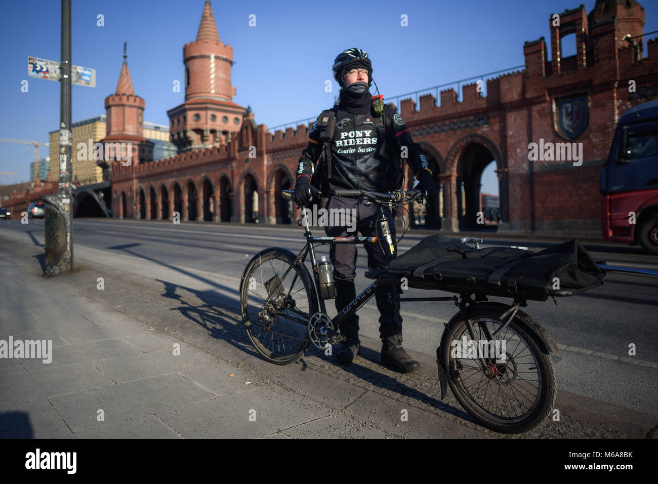 28 February 2018, Germany, Berlin: Bike courier Anselm Holthaus sitting on  his bike with a neck scarf wrapped around him on the Oberbaum bridge in  Kreuzberg in ice-cold temperatures. Due to the