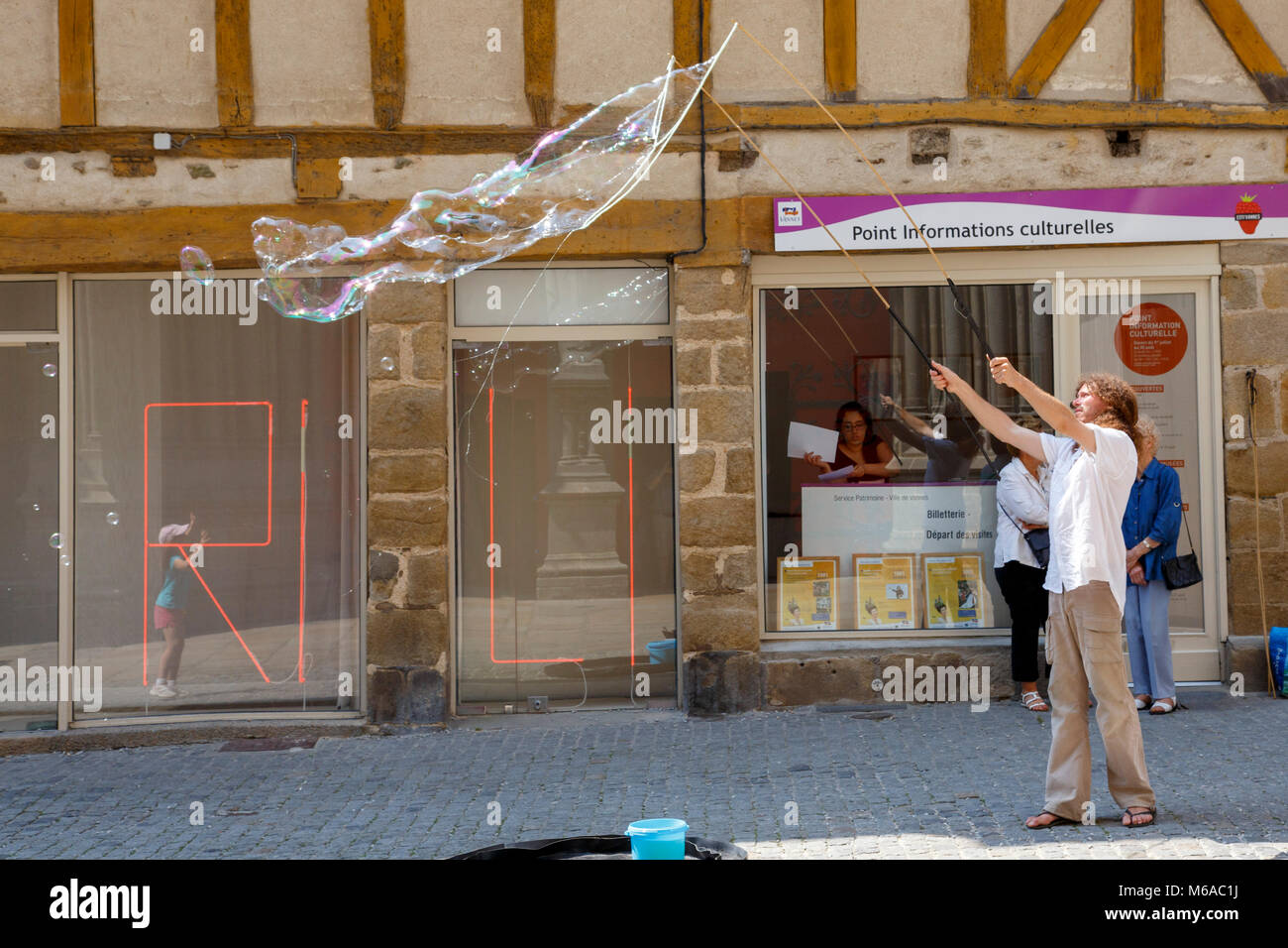 Street entertainer makes soap bubbles.  The reflection of a young girl can be seen in the window waiting to burst the bubbles as they float past her. Stock Photo