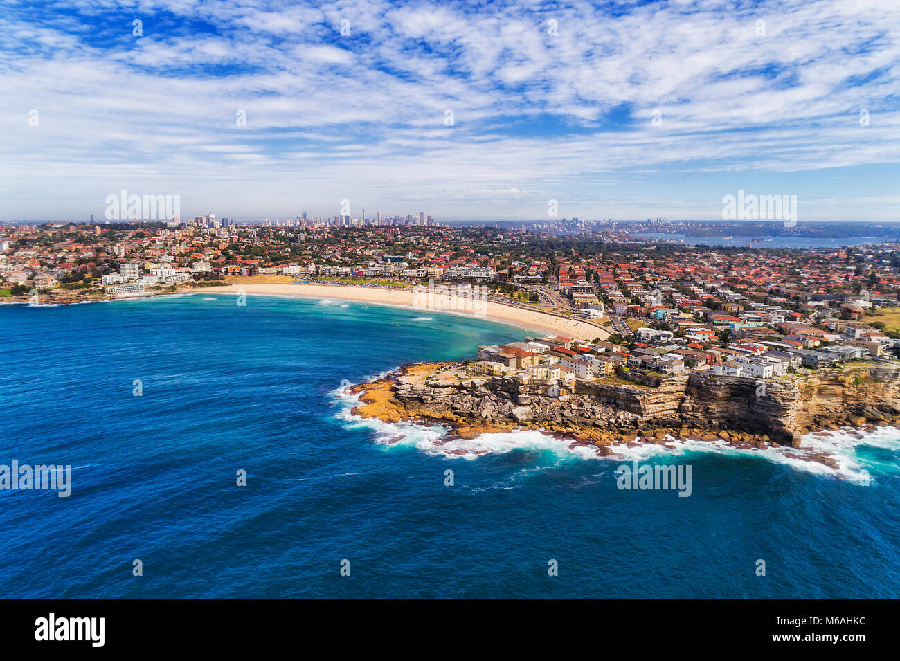 Elevated aerial view from open sea to North Bondi cliff head, sand of Bondi beach and distant city CBD towers on horizon. Stock Photo