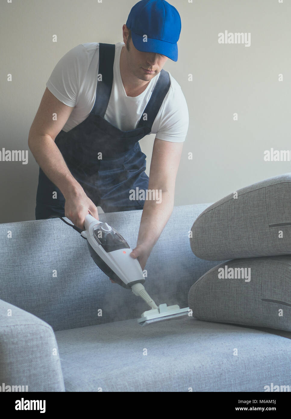 Man in uniform cleaning fabric of the sofa with dry steam cleaner Stock