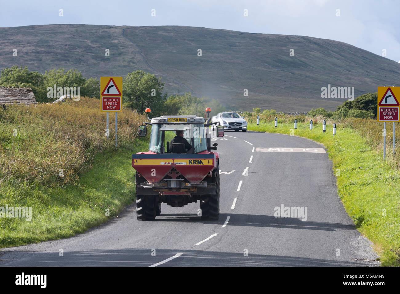 Rear view of tractor pulling fertiliser spreader & front of Mercedes convertible driving & travelling on country road - North Yorkshire, England, UK. Stock Photo