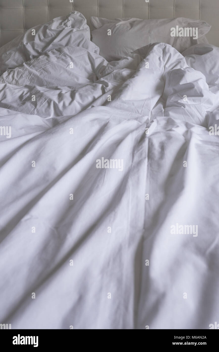 Unmade bed with plain white bed linen in a close up view of a rumpled duvet, pillows and a padded headboard Stock Photo