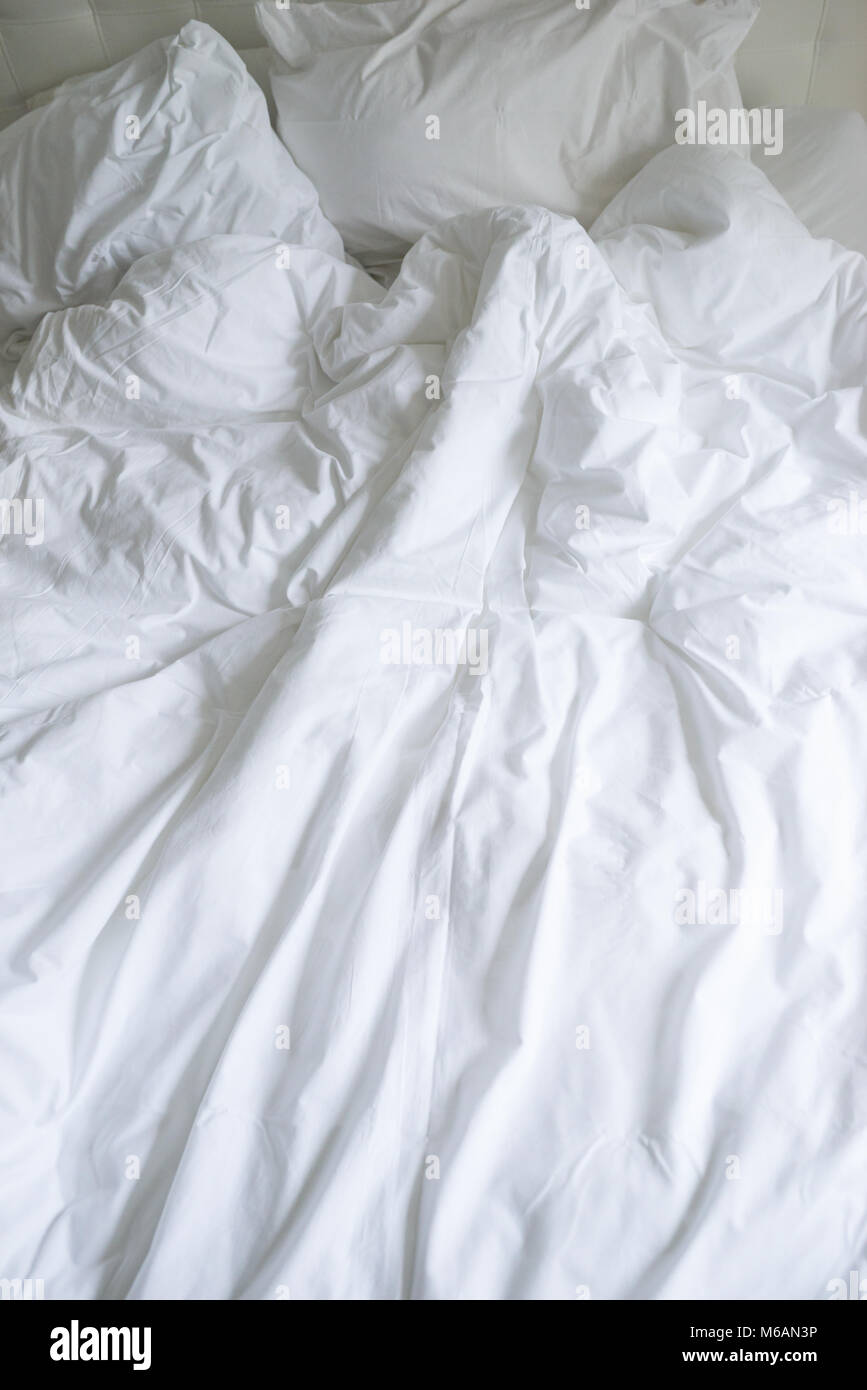 Unmade bed with plain white bed linen in a close up view of a rumpled duvet, pillows and a padded headboard Stock Photo