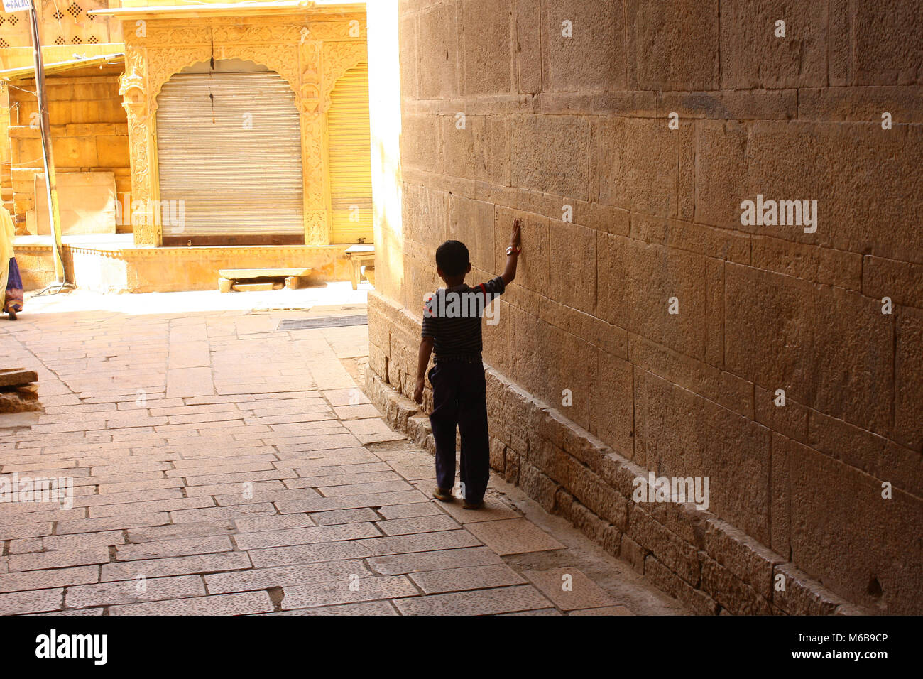 Black silhouette of a little boy walking through an archway into a sunny street, sweeping with one hand along the massive stone wall. Rajastan, India. Stock Photo