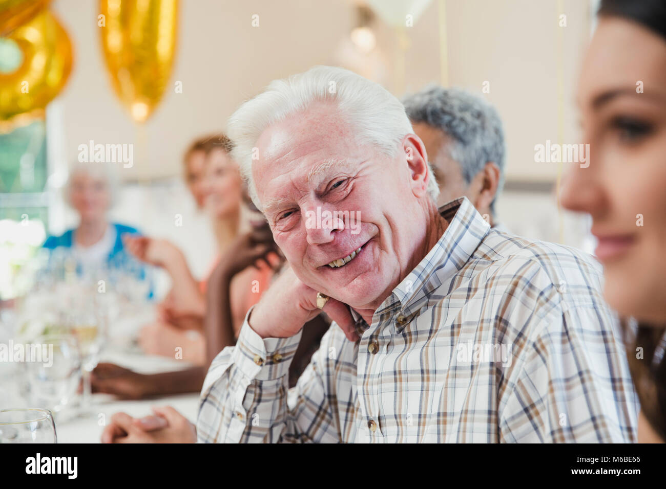 Happy senior man at a birthday party with his friends and family. He is sitting at the table smiling for the camera with his head resting on his hand. Stock Photo