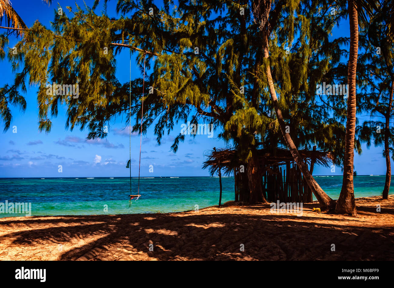 A wooden hut on the sand and a rope swing under the shade of trees and coconut palms on the shore of the blue ocean with white waves, sky and clouds o Stock Photo