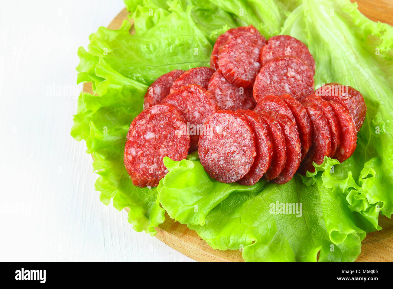 Smoked sausage, salami chopped in slices on a salad on a wooden circular cutting board on a white table Stock Photo