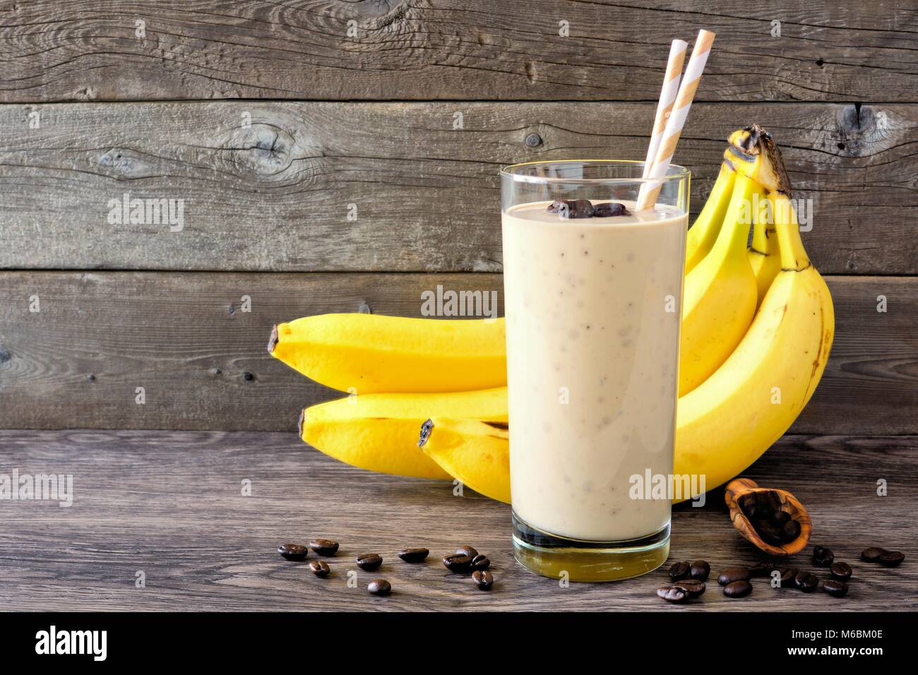 Coffee, banana smoothie in a tall glass with coffee beans and bananas in background. Side view, against a rustic wood background. Stock Photo