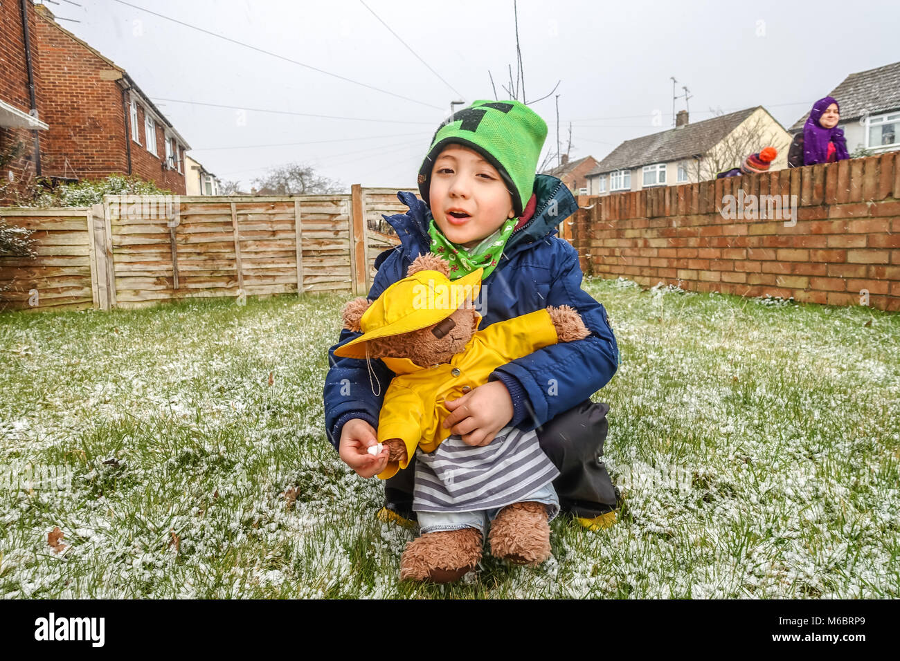 A young boy plays with his teddy bear in the front garden in the snow. Both the child and the toy are dressed in winter hats and coats. Stock Photo