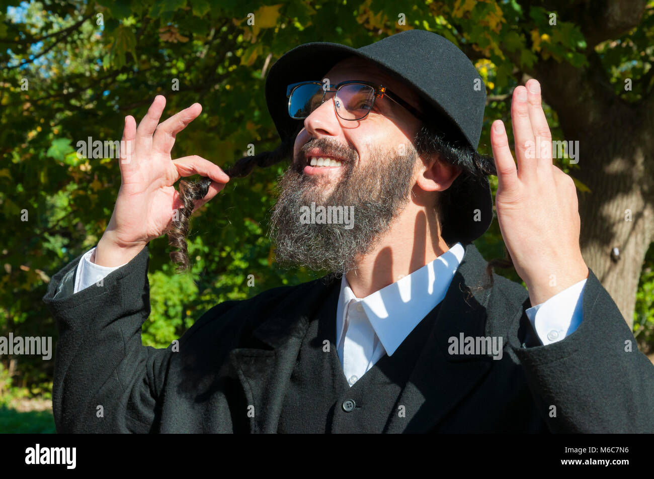 Outdoor sunny portrait of a young happy orthodox Jewish man with eyeglasses and black beard holding payot, sidelocks and looking up to the sky Stock Photo
