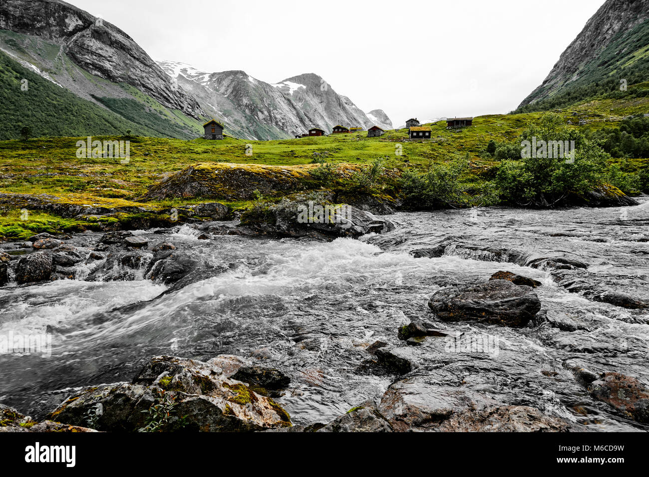 Beautiful river with clear water and a rocky shore. Colorful houses on green grass and snow capped mountains in the background in Norway. Stock Photo