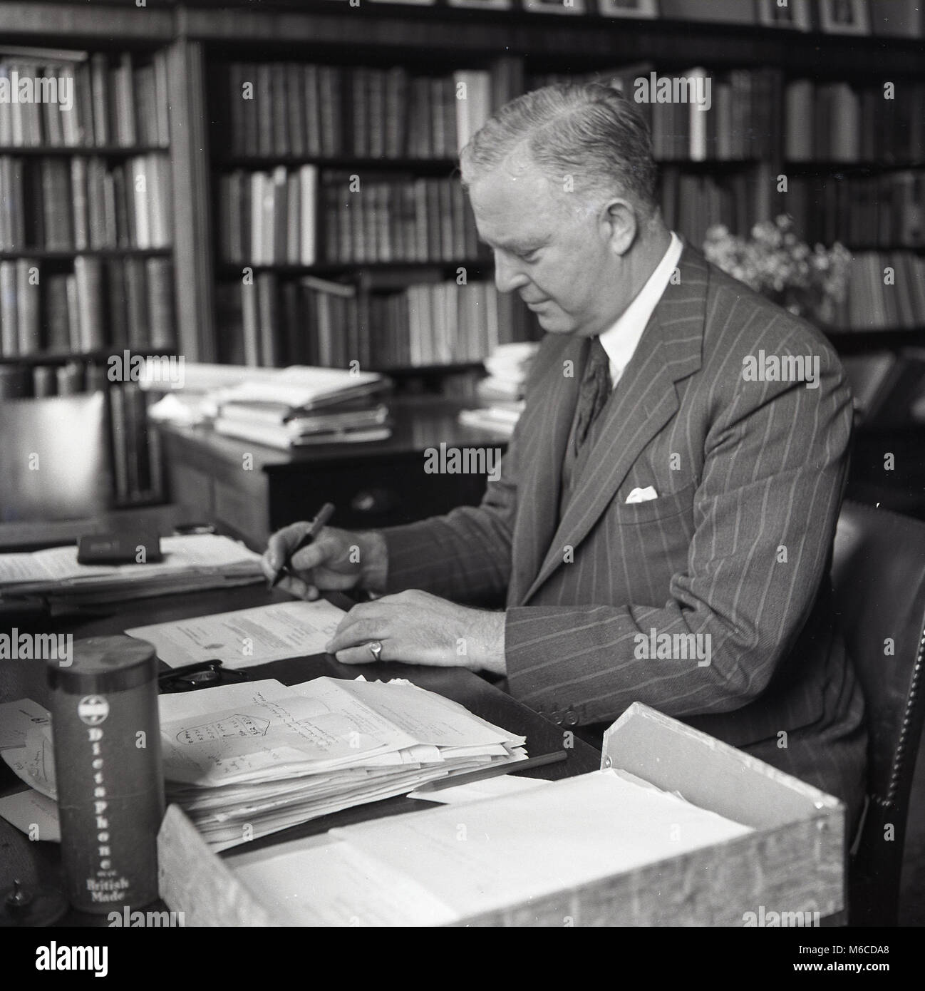 1950s, historical, a well-dressed English gentleman in a pin-stripe suit sitting at a desk in a book filled study, reading correspondence and signing letters, England, UK. Stock Photo