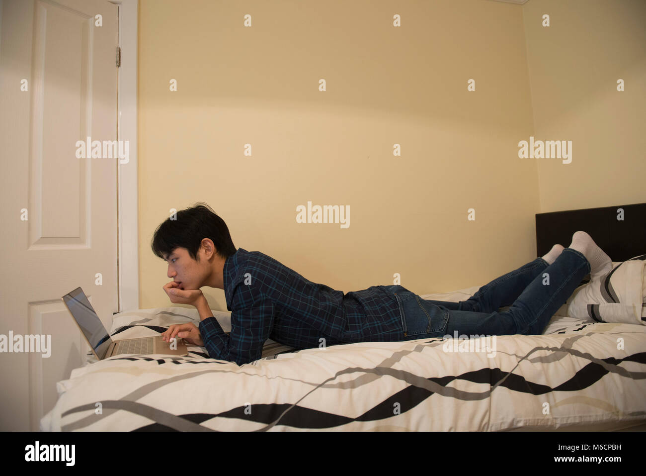 a young asian male student relaxes on his bed while working on his ...