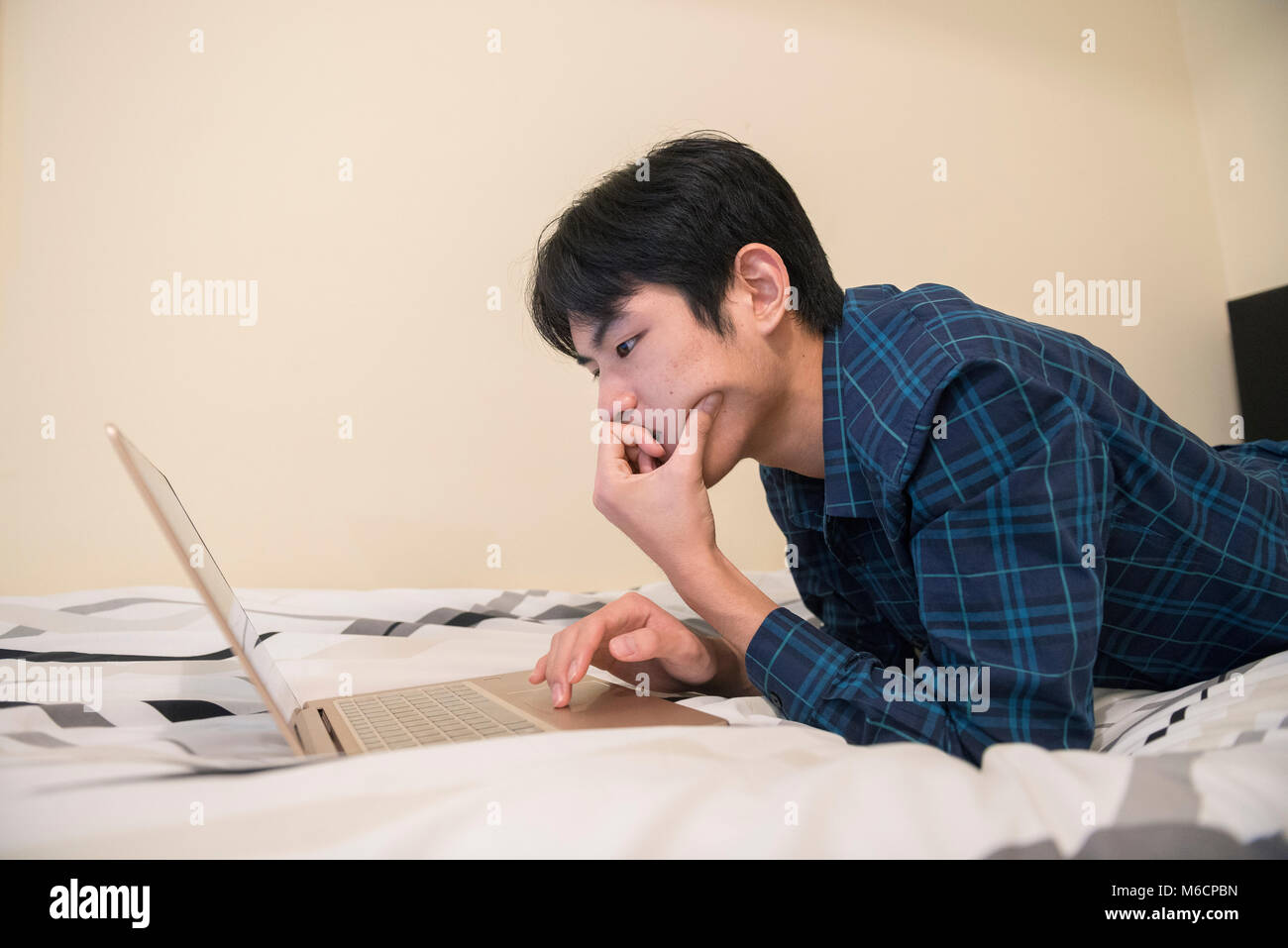 a young asian male student relaxes on his bed while working on his ...