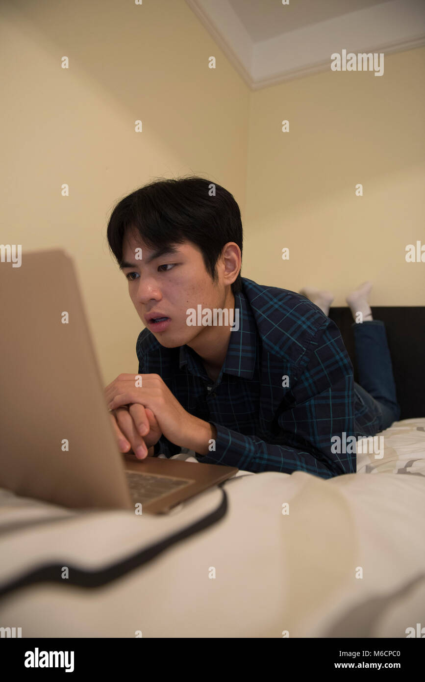 a young asian male student relaxes on his bed while working on his ...