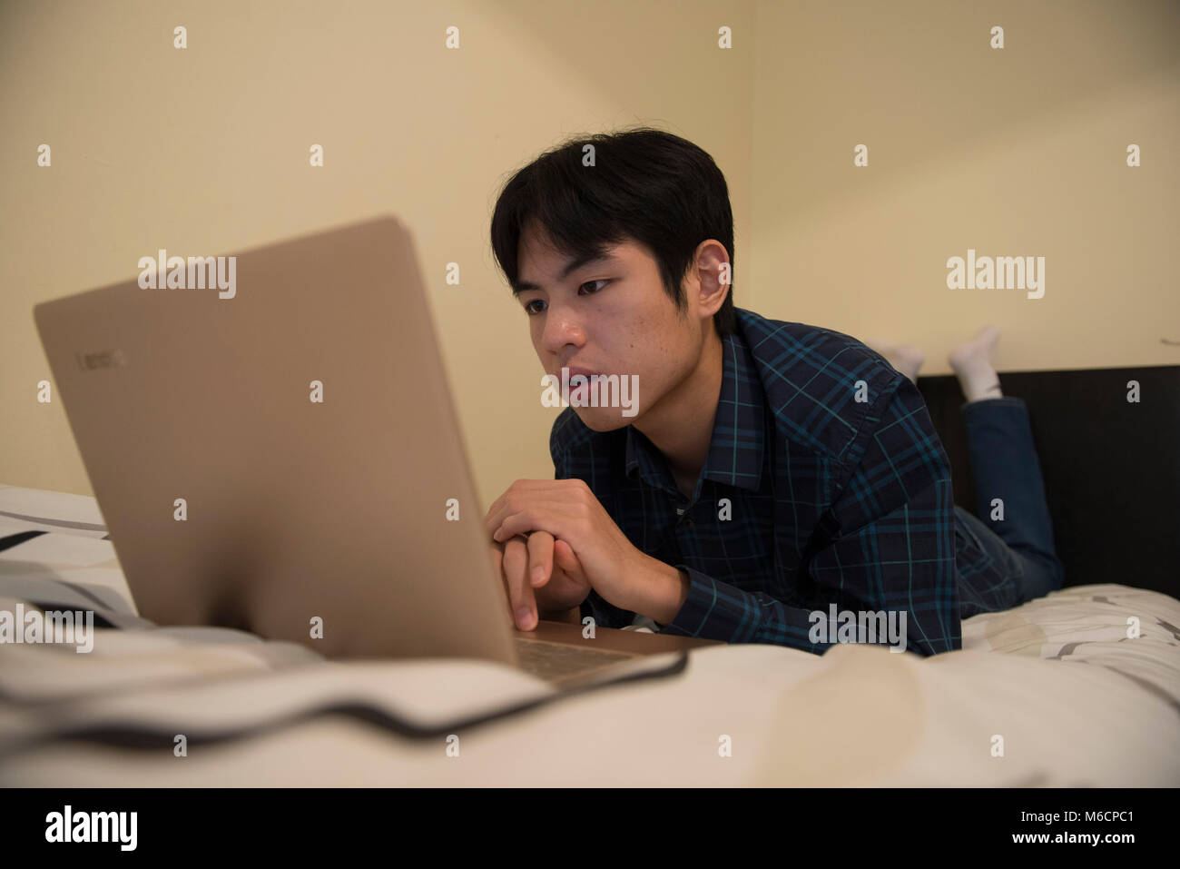 a young asian male student relaxes on his bed while working on his ...