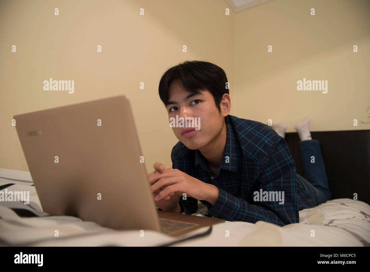 a young asian male student relaxes on his bed while working on his ...