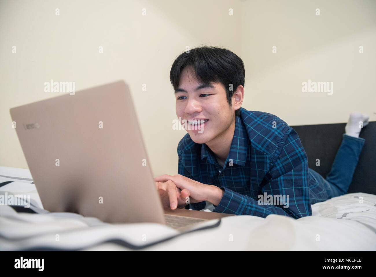 a young asian male student relaxes on his bed while working on his ...