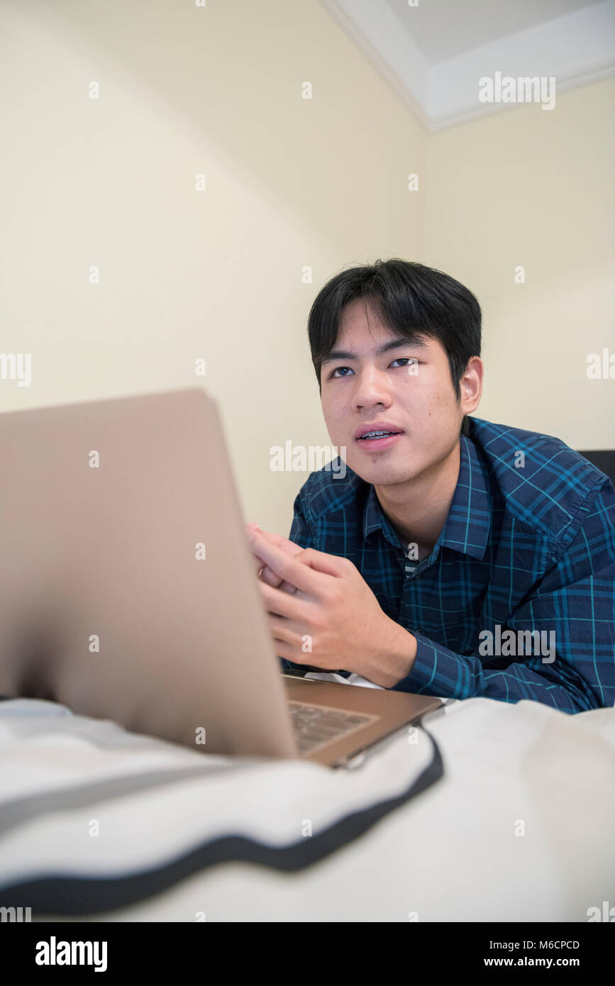 a young asian male student relaxes on his bed while working on his ...