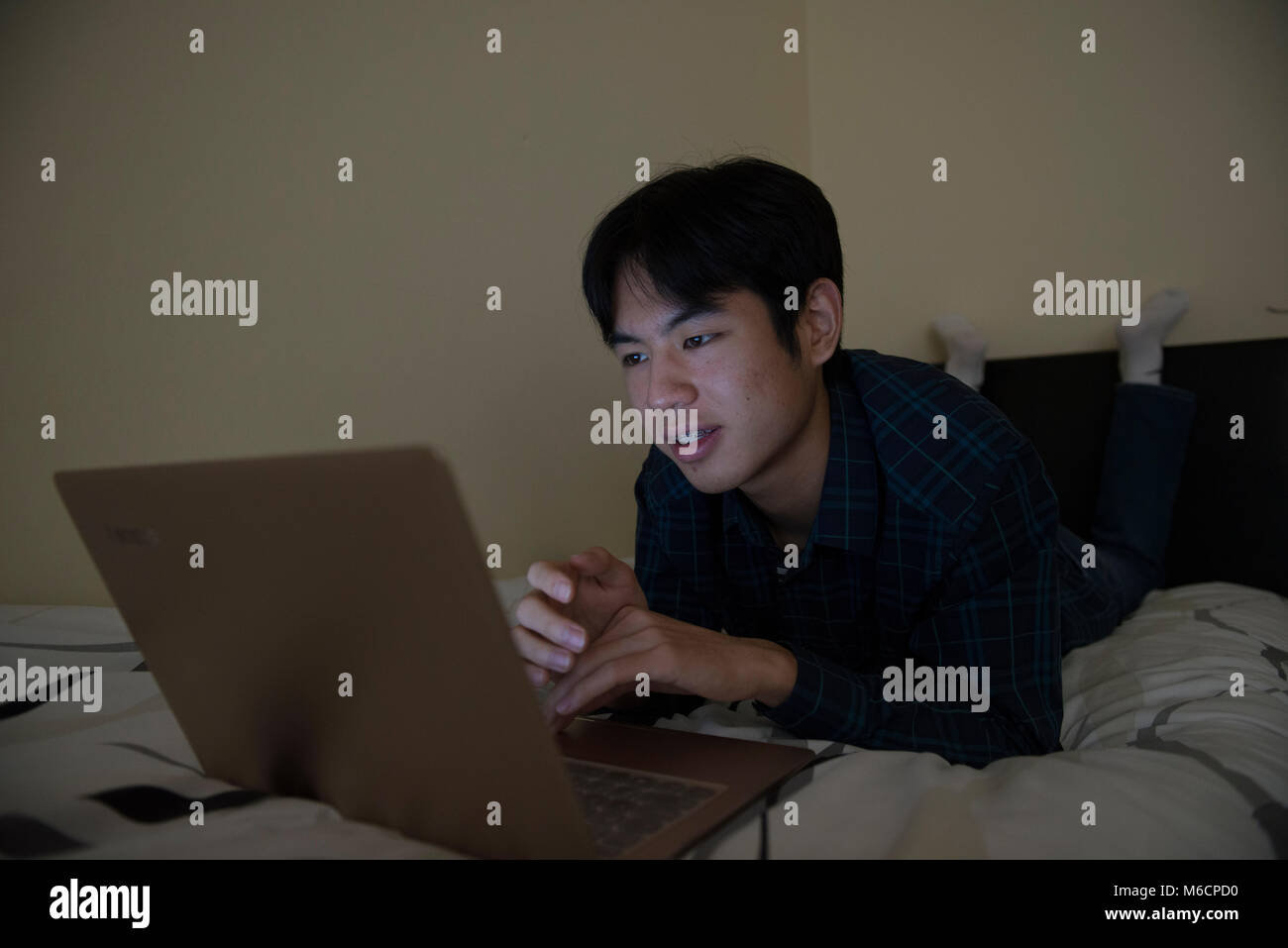 a young asian male student relaxes on his bed while working on his ...