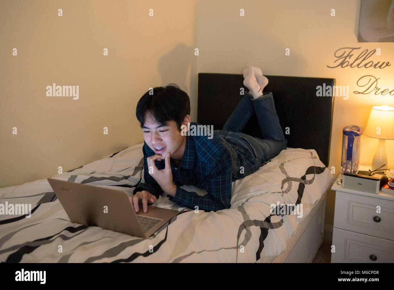 a young asian male student relaxes on his bed while working on his ...