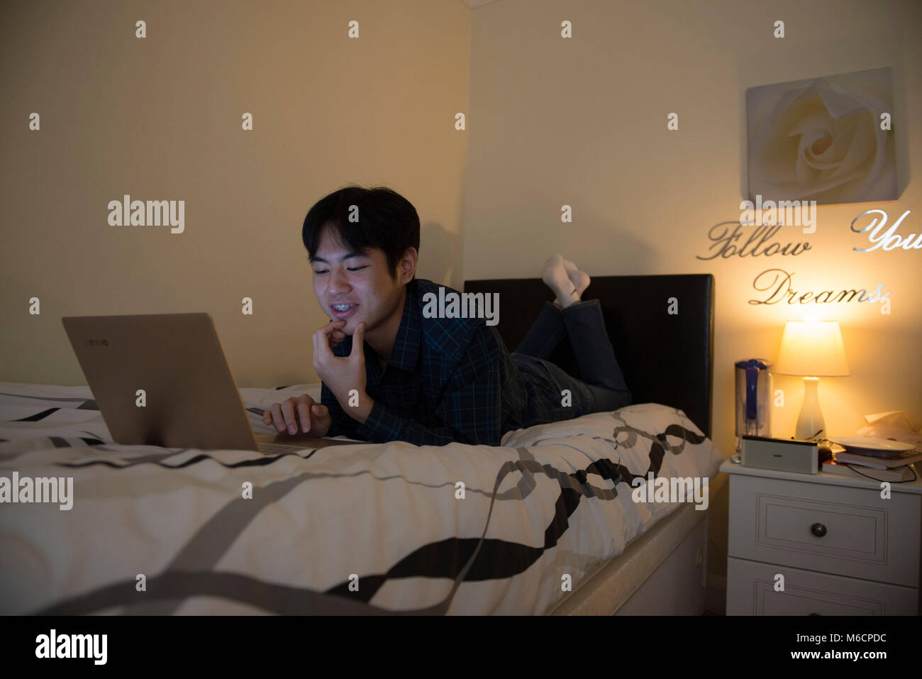 a young asian male student relaxes on his bed while working on his ...