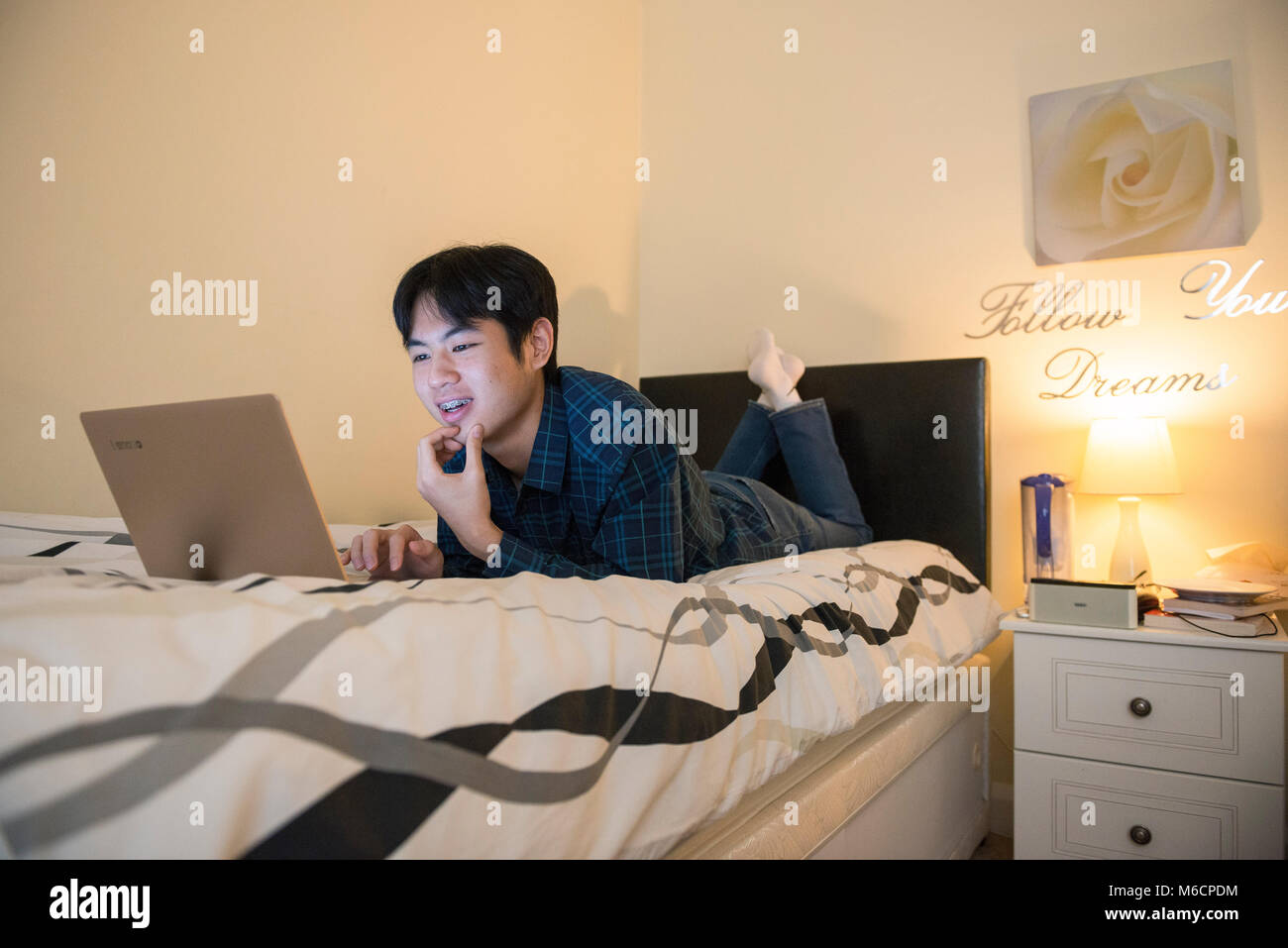 a young asian male student relaxes on his bed while working on his ...