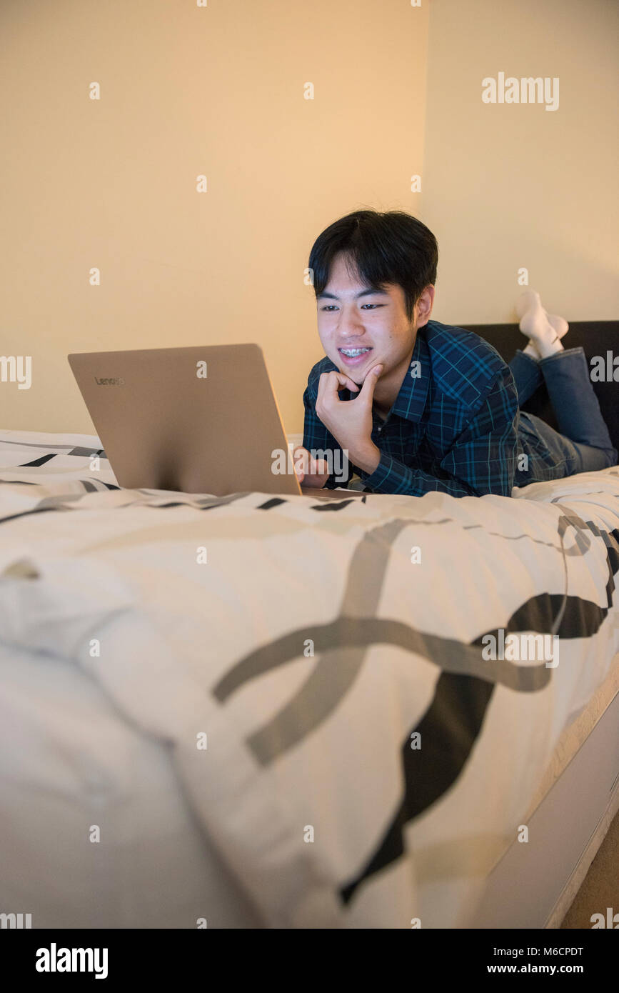 a young asian male student relaxes on his bed while working on his ...