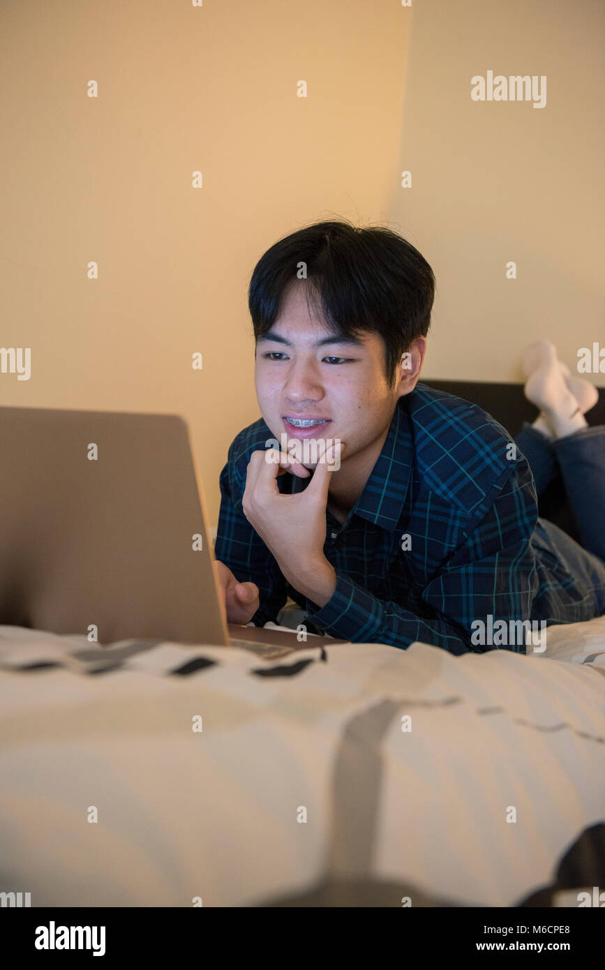 a young asian male student relaxes on his bed while working on his ...