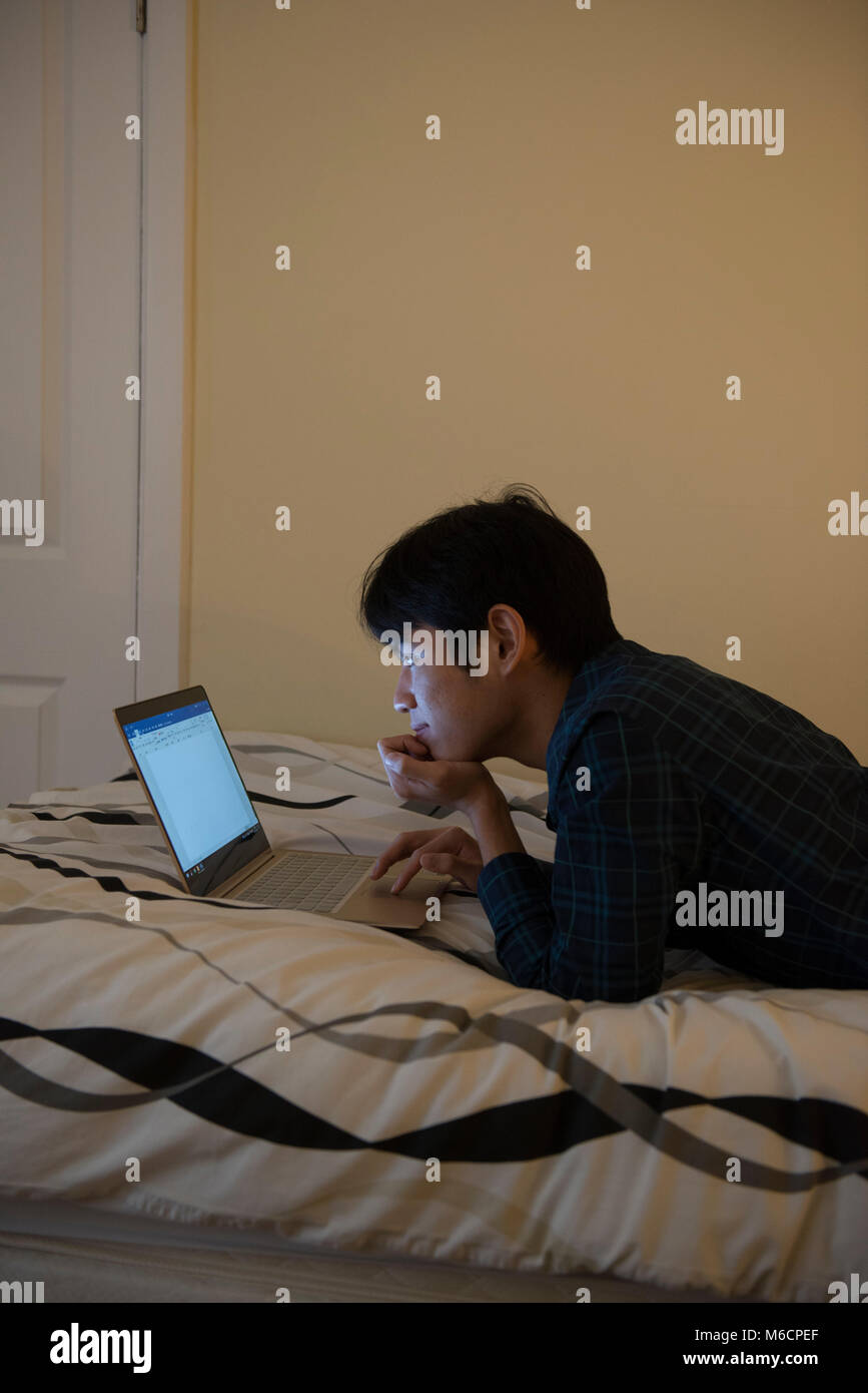 a young asian male student relaxes on his bed while working on his ...