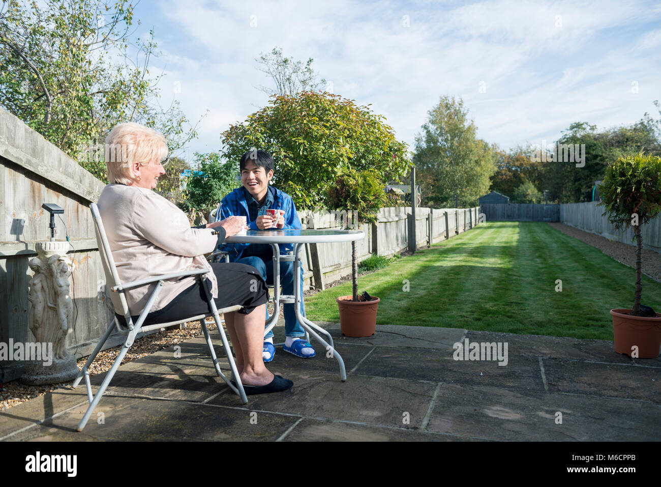 a student accommodation host sits with her international student in the garden drinking tea and talking. Stock Photo