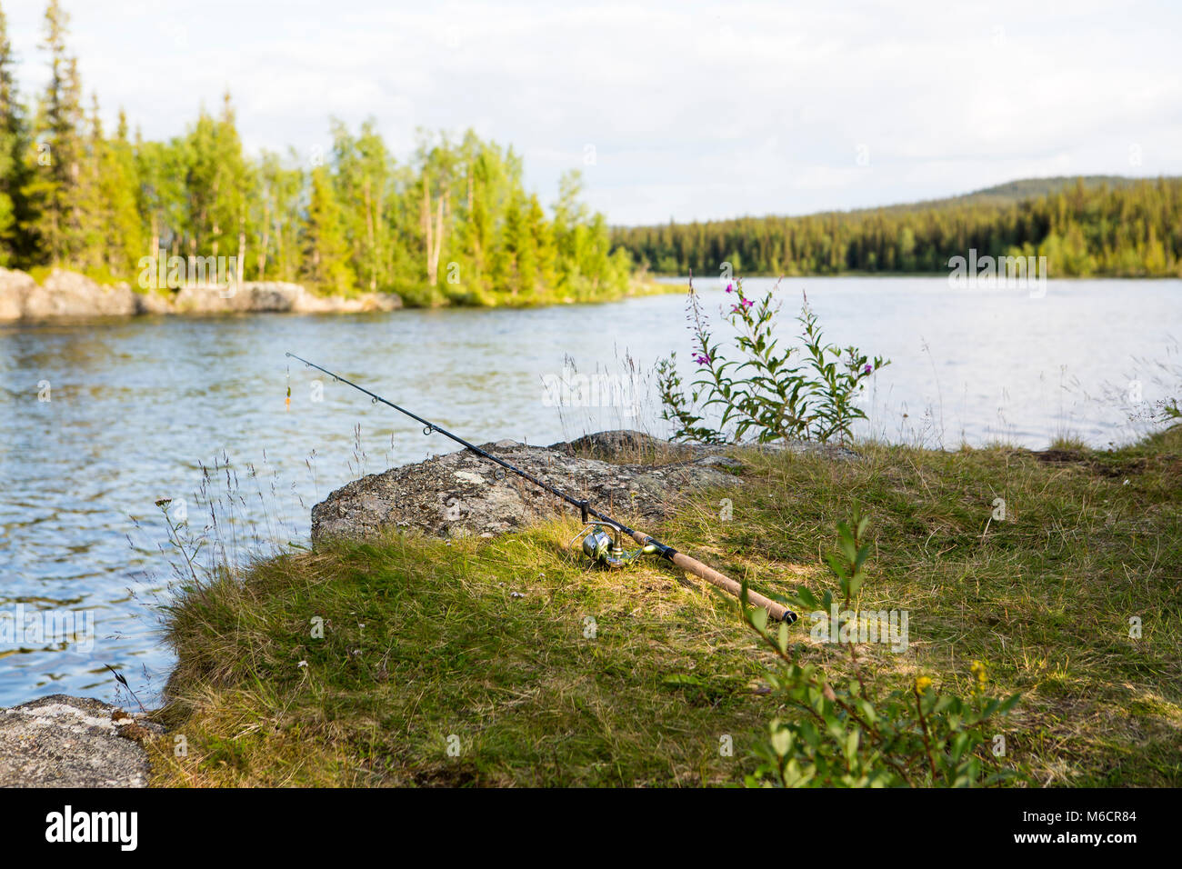 Fishermans fishing rod and reel placed on the ground by the water. A wild untouched nature is in the background. Stock Photo