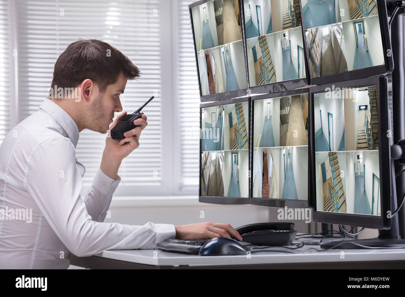Young Male Security Guard Talking On Walkie-talkie While Monitoring Multiple CCTV Footage On Computer Stock Photo