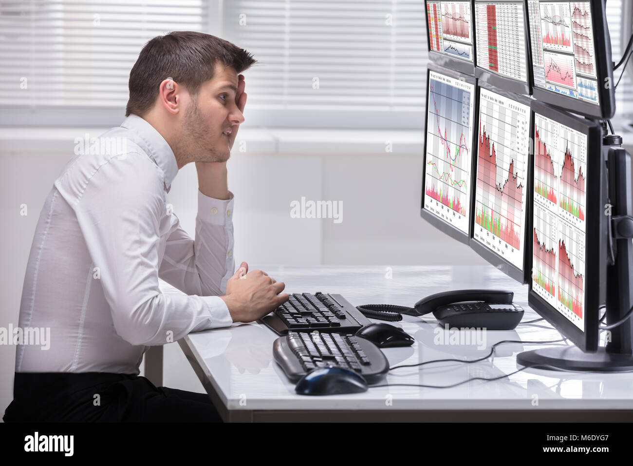 Side View Of A Stressed Young Male Operator Looking At Graphs On Multiple Computer Screens Stock Photo