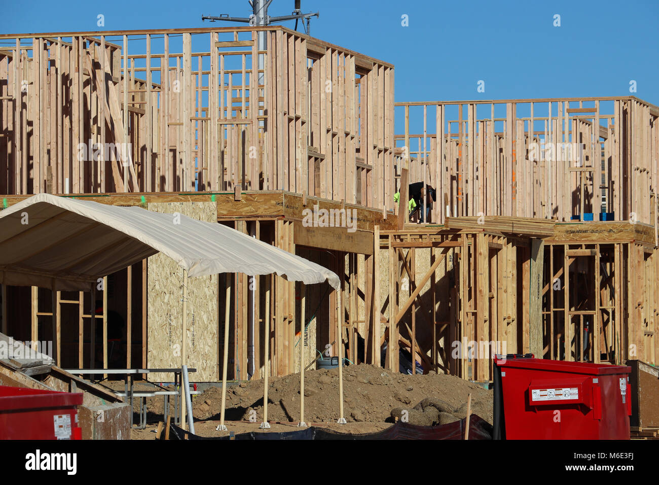 Several new two-story homes under construction, just framed, with skeletal walls built of 2 x 4 lumber; blue sky, red dumpster, shade, on sunny day. Stock Photo