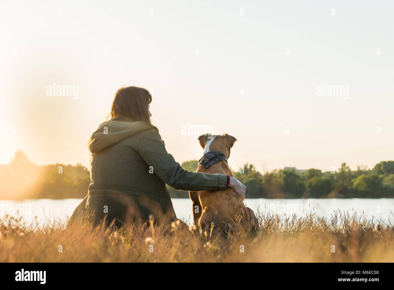 Dog owner and her pet sit at riverbank at sunset Stock Photo
