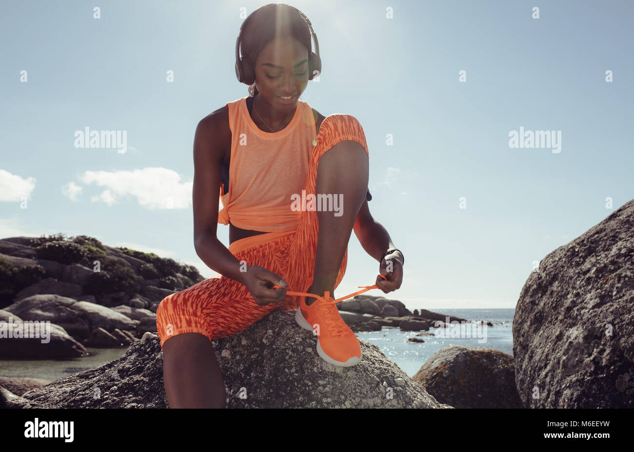 Fit and sporty young woman tying her laces before a run. Female runner tying her shoelaces while training at the rocky beach. Stock Photo